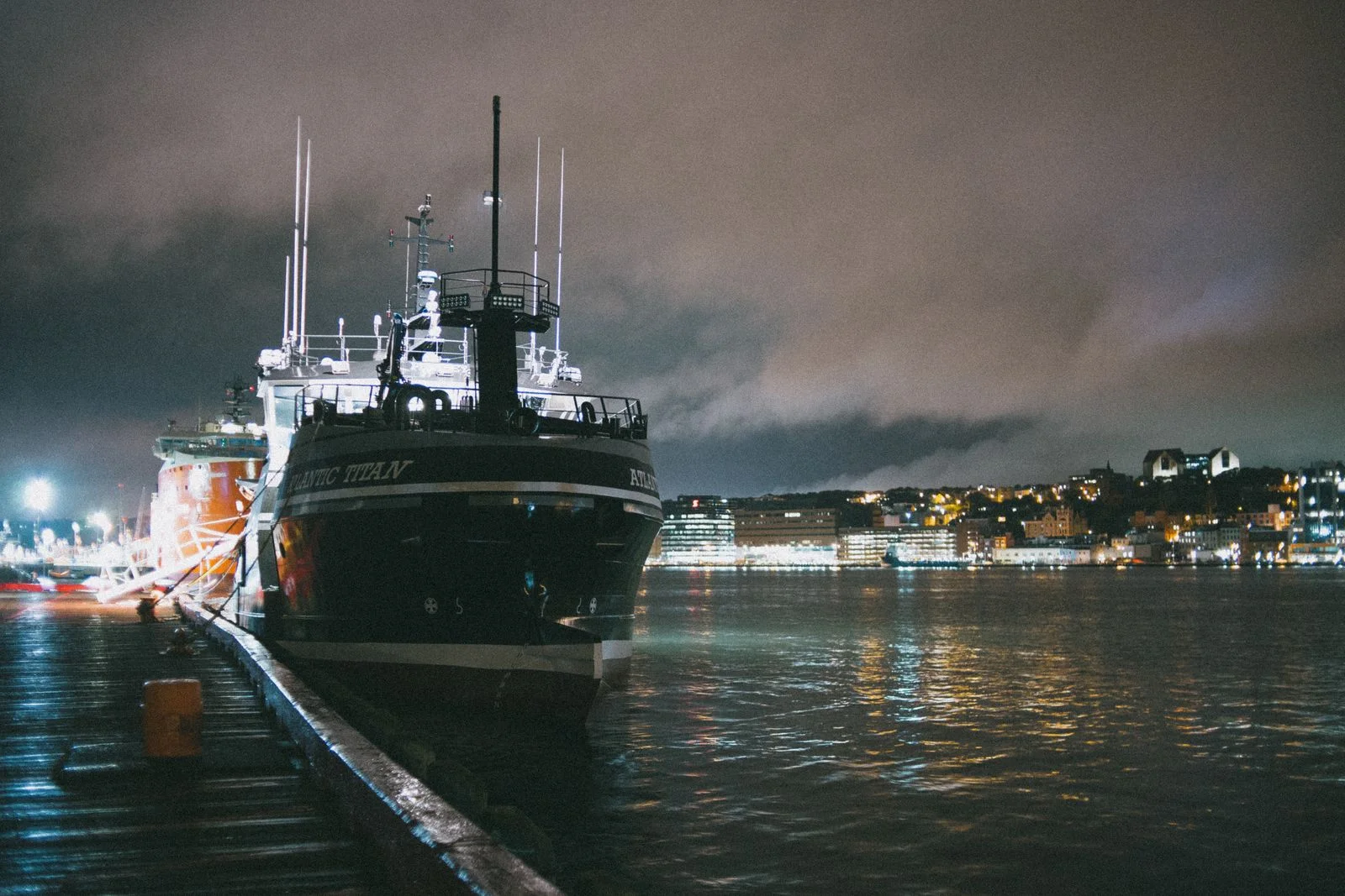 A large ship moored at a city quay at night, lights reflected in the water